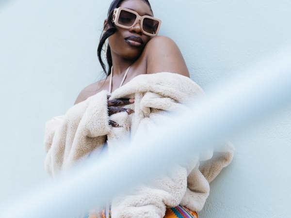 Frau mit großer Sonnenbrille und bunter Hose lehnt an einer Wand